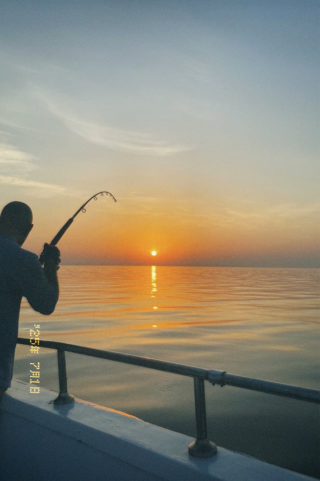 Someone holding a bent fishing rod on a boat as the sun sits low over still water.